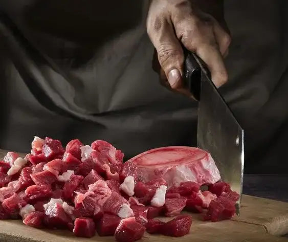 En skarp, sort-håndteret hakkeøks bruges til at hakke rå rødt kød i små tern på et træskærebræt.