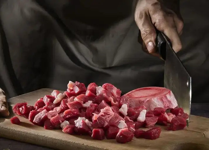 Et rustikt træskærebræt holder en bunke frisk ternet rødt kød med synligt hvidt fedt, sammen med et delvist skåret stykke med knogler.